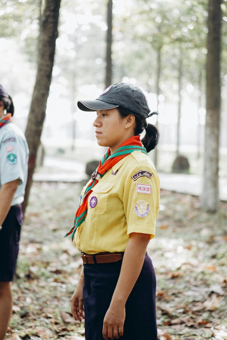 Scout leader wearing uniform stands in a forest setting during a group event.